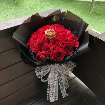 A bouquet of vibrant red roses with a golden rose at its center, wrapped in sleek black paper and adorned with a large white lace ribbon.