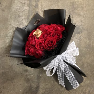 A lush bouquet of deep red roses with a single golden rose in the middle, arranged in matte black wrapping paper and tied with a white lace bow.