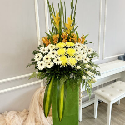 funeral flower on a tall stand featuring yellow chrysanthemum, white gerbera, white rose and orange crab with orchid in a layered arrangement