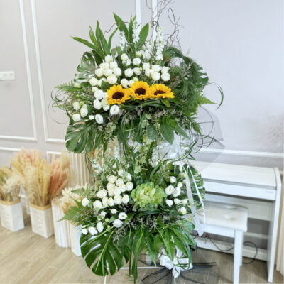flower stand for funeral event featuring white rose, white eustoma, and sunflower with lush green leaves and blue caspia accents