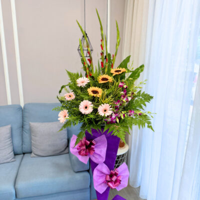 opening flower stand featuring gladiolus, sunflower, pink gerbera and orchid with lush green foliage wrapped in purple paper