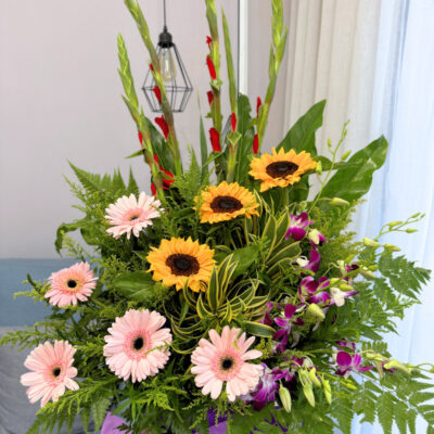 a vibrant congratulation flower stand with tall gladiolus, bright sunflower, soft pink gerbera and delicate orchid in purple wrap