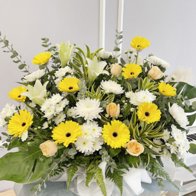 a floral tribute stand with bright yellow gerbera, white chrysanthemum pompom, madonna lily and rose arranged in a tall display