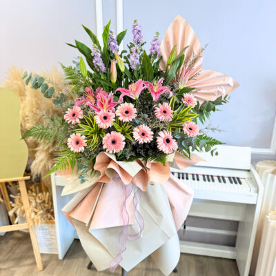 a tall flower stand with a mix of pink gerberas, stargazer lilies, and purple matthiola wrapped in layered pastel paper.