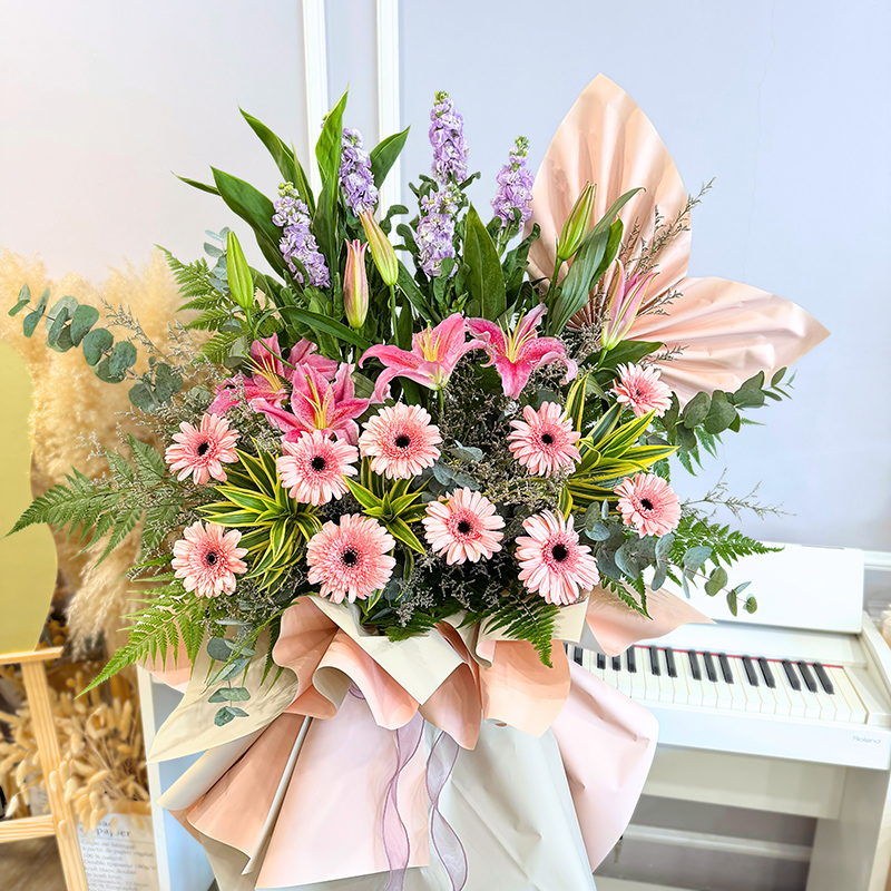 mixed flower bouquet stand including pink gerberas, stargazer lilies, and matthiola, styled with leafy greens and peach-toned paper wrap.