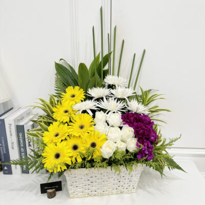 a funeral flower basket featuring white chrysanthemum, yellow gerbera, white rose and purple carnation with tall green foliage