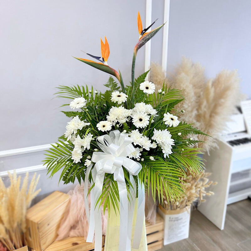 flower stand featuring bird of paradise, white gerbera, and white chrysanthemum pompom with green leaves and white ribbon