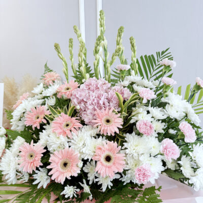 a tall funeral stand featuring mixed flowers including pink carnations, white chrysanthemum pompoms and hydrangeas arranged with green foliage