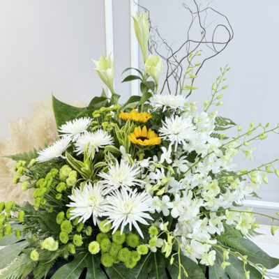 a funeral flower with tall madonna lilies, white and green chrysanthemums, sunflowers and white orchids arranged on a black stand
