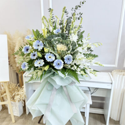 condolences flower stand featuring blue gerbera, white orchid, tuberose and white brassica arranged on a funeral stand wrapped in soft mint green paper.