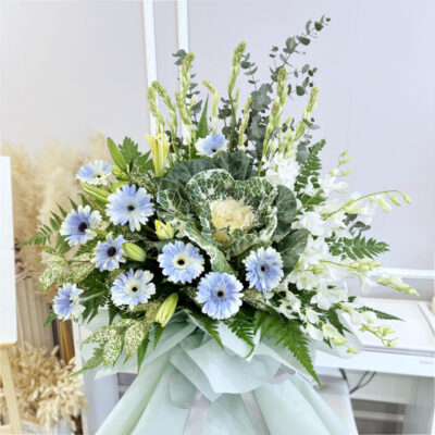 a flower stand with delicate white orchid, blue gerbera, tuberose and white brassica, wrapped in pale green material.