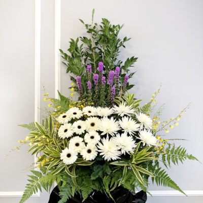 a tall funeral flower stand featuring white chrysanthemum, white gerbera, purple liatris, and yellow dancing lady flowers on a black-wrapped stand