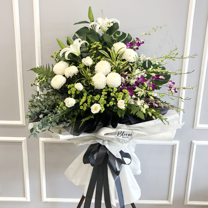 flower stand of white Casablanca lilies, chrysanthemums, roses, pompom chrysanthemums and purple orchids arranged on a tall funeral stand wrapped in white and black paper.