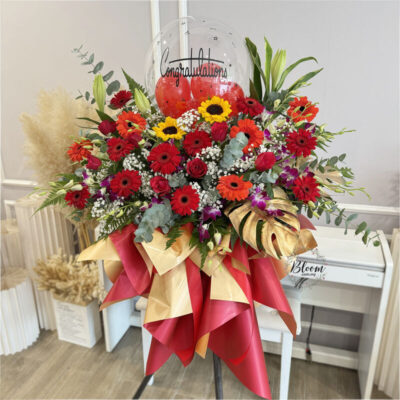 opening flower stand featuring red gerbera, red rose, sunflower, orchid and baby's breath with a congratulation balloon