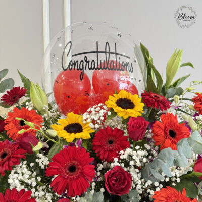 a vibrant congratulations flower stand with red gerbera, sunflower, red rose, orchid and baby's breath topped with a clear balloon