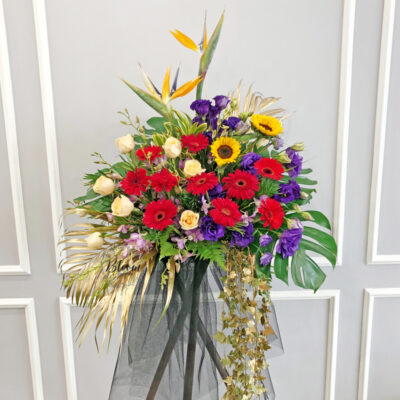 a tall wooden flower stand featuring a mix of tropical bird of paradise, red gerberas, champagne roses, orchids, sunflowers and purple eustomas