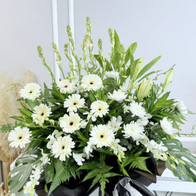 a funeral flower stand featuring white lilies, orchids, gerbera and chrysanthemum pompoms on a black wrapped stand
