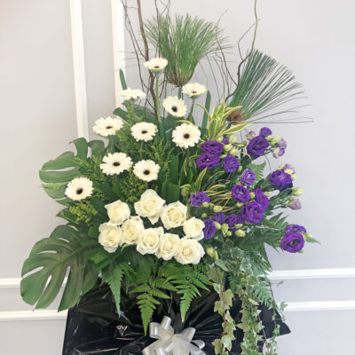 funeral floral stand with white gerbera, white roses, purple eustoma and green foliage tied with a silver ribbon on black wrapping
