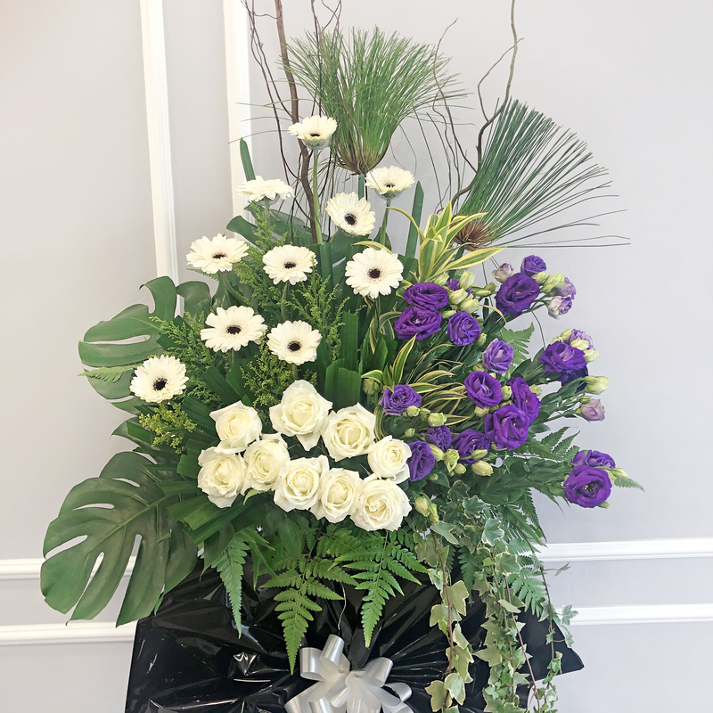 funeral floral stand with white gerbera, white roses, purple eustoma and green foliage tied with a silver ribbon on black wrapping