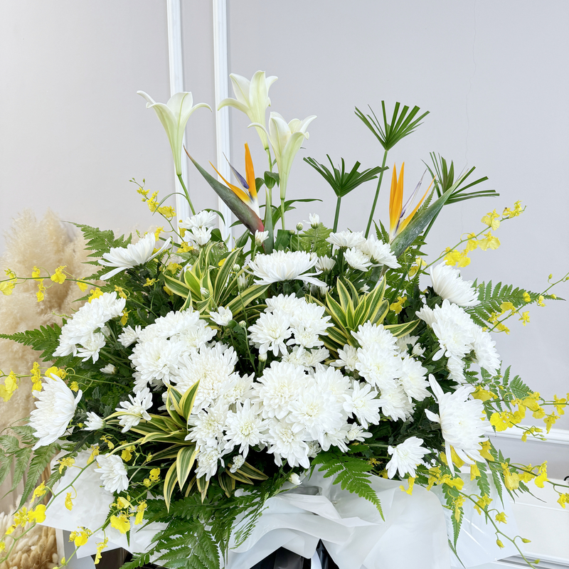 a tall condolences flower stand featuring madonna lily, bird of paradise and white chrysanthemum with yellow dancing lady accents wrapped in black