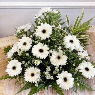 a condolences basket flower arranged with white gerbera daisies and white chrysanthemum pompom surrounded by lush green leaves