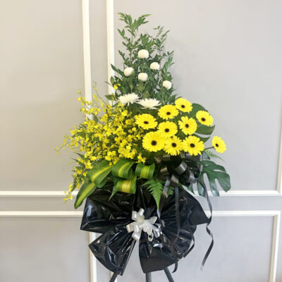 a funeral flower stand featuring white chrysanthemum ping pong, white chrysanthemum, yellow gerbera and dancing lady orchids arranged on a tall stand with lush green foliage