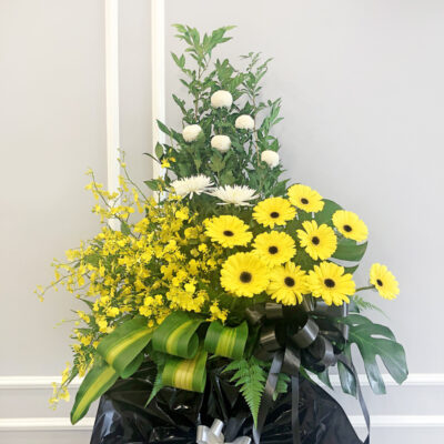condolences flower stand arranged on a tall stand with white chrysanthemum ping pong, yellow gerbera, white chrysanthemum and delicate dancing lady orchids surrounded by green leaves