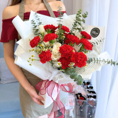 flower bouquet featuring vibrant red carnations, white eustoma, and chamomile wrapped in white paper with a sheer red ribbon