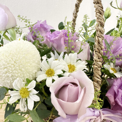 a basket flower arrangement with delicate lilac rose, purple eustoma, white chrysanthemum ping pong and white hana flower in natural basket