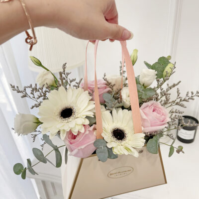 a flower arrangement styled in a handbag box featuring pink roses, white gerberas, and mixed greenery with delicate filler flowers