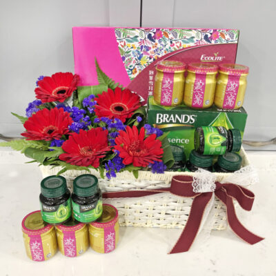 basket arrangement featuring red gerbera flowers with purple fillers alongside bottles of bird's nest and essence of chicken in a white woven basket with a maroon ribbon.