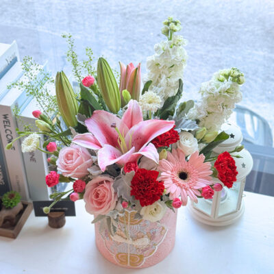 flower arrangement in a decorative box featuring pink stargazer lilies, pink roses, red carnations and white matthiola