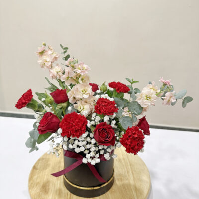 flower arrangement featuring red rose spray, red carnations, soft matthiola, and baby's breath arranged in a round black box with a maroon ribbon