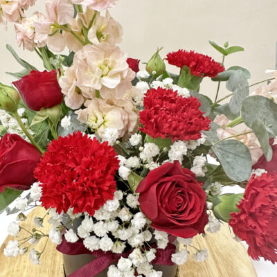 a flower arrangement with vibrant red carnations and roses, complemented by pale pink matthiola and white baby's breath in a dark box