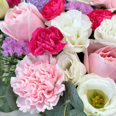 a flower arrangement in a round pink box with pink carnations, pink roses, carnation spray, and purple chrysanthemum pingpong flowers