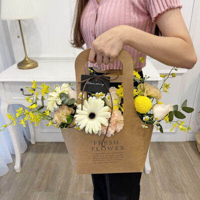 flower arrangement in a brown handbag box featuring champagne roses, white gerberas, and yellow chrysanthemums