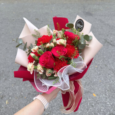 flower bouquet wrapped in red and cream paper featuring red roses, red carnations, and light carnation sprays with greenery.