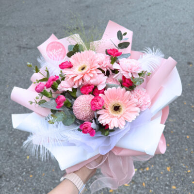 flower bouquet with pink gerbera, pink carnations, pink chrysanthemum ping pong, and white feather accents wrapped in pastel paper.
