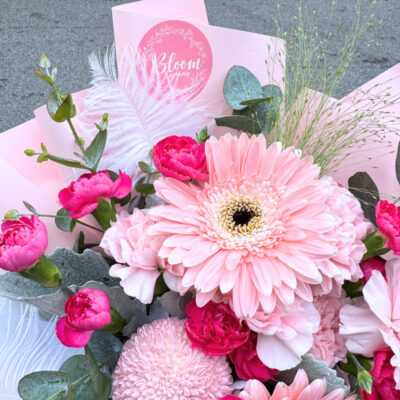 a bouquet featuring pink carnations, gerbera, chrysanthemum ping pong, and delicate feathers arranged with pink and white wrapping.