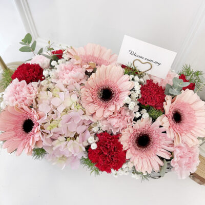 a arrangement in a woven basket with pink gerbera, soft pink hydrangea, and red carnations accented by white filler flowers