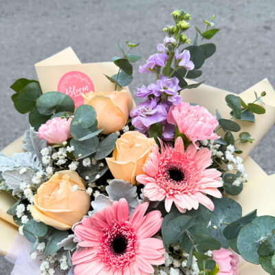 a bouquet featuring pink carnation, champagne rose, pink gerbera, and delicate greenery with soft cream wrapping