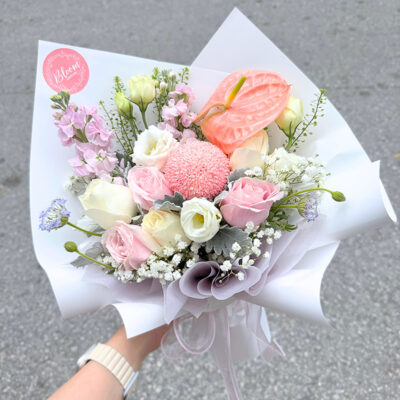 a flower bouquet wrapped in lilac paper featuring pink matthiola, pink and white roses, pink anthurium, and white eustoma with baby’s breath accents