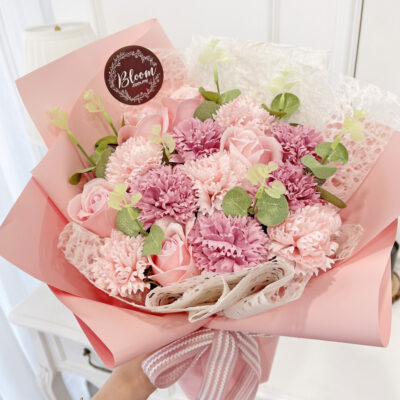 a flower bouquet featuring pink soap carnations and pink soap roses wrapped in soft pink paper with white lace accents