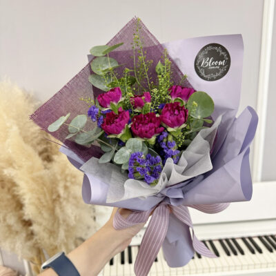 hand bouquet with six purple carnations wrapped in lilac and white paper accented with eucalyptus and purple fillers