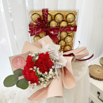 flower bouquet with three red carnations and white filler flowers wrapped in soft pink paper, paired with a box of ferrero rocher chocolates tied with red ribbon