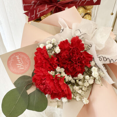 a small bouquet featuring vibrant red carnations and delicate white blooms wrapped in pink paper alongside a clear box of ferrero rocher chocolates