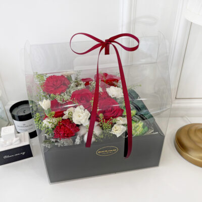 flower arrangement in a clear box with red roses, red carnations, white spray carnations, and cotton flowers tied with a red ribbon
