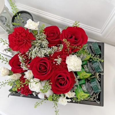 a flower arrangement featuring red roses and carnations with white spray carnations and cotton flowers inside a transparent gift box