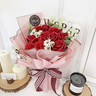 a flower bouquet of red soap carnations and red soap roses wrapped in pink translucent paper with printed newspaper style paper and tied with a striped ribbon