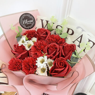 wrapped bouquet featuring red soap roses and carnations accented with small white soap flowers and greenery in a soft pink and printed paper wrap